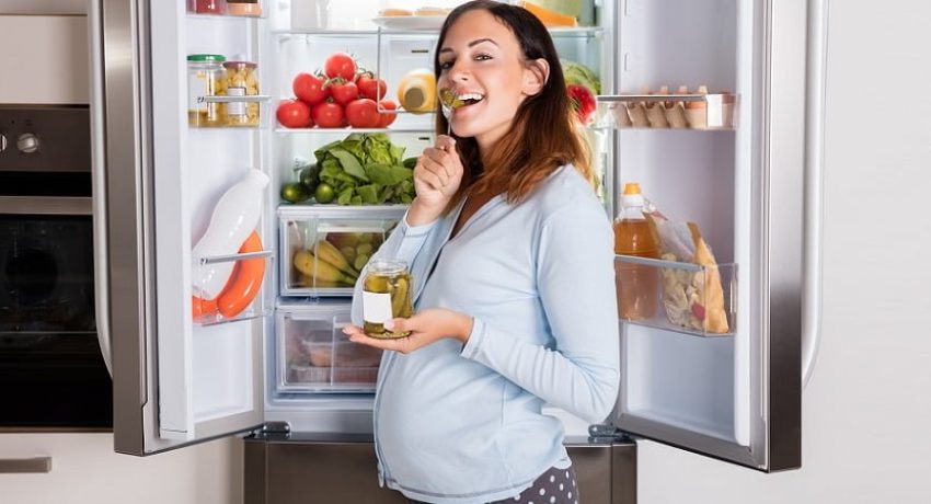 Young Pregnant Woman Enjoy Eating Jar Of Pickle In Front Of Open Refrigerator In Kitchen