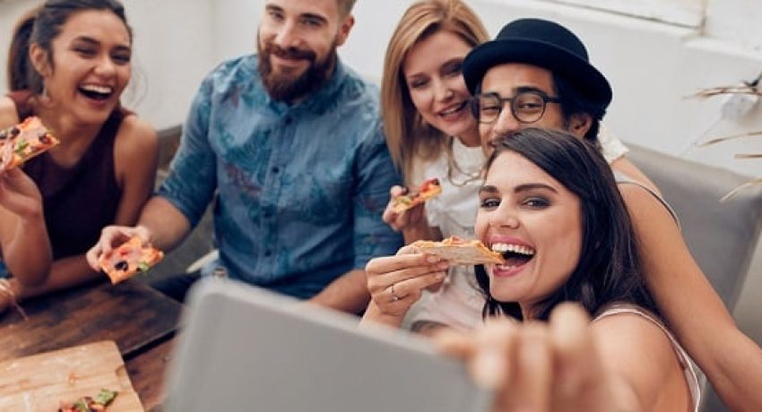 Group of multiracial young people taking a selfie while eating pizza. Young woman eating pizza her friends sitting around during a party.