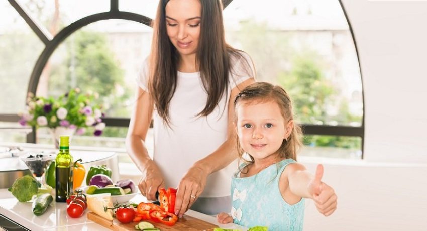 Cute little girl cooking with her mother and shows thumbs up. Healthy food, cooking healthy salad with vegetables ingredients. Mom and daughter cooking together. Recipe food for baby or child